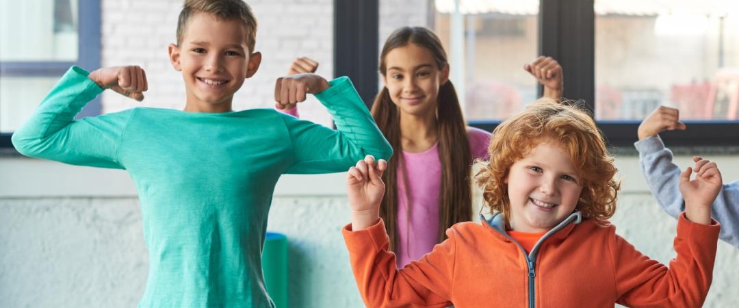 Four smiling children in colorful clothes strike fun muscle poses indoors, conveying joy and confidence. Bright window light in the background.