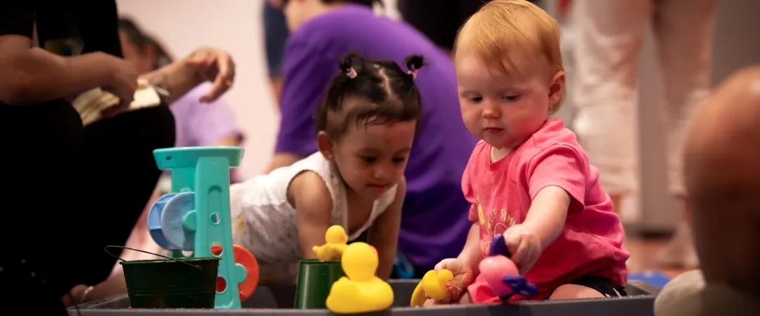 Two curious toddlers play with colorful bath toys at a sensory water table as adults supervise, creating a lively, playful scene.