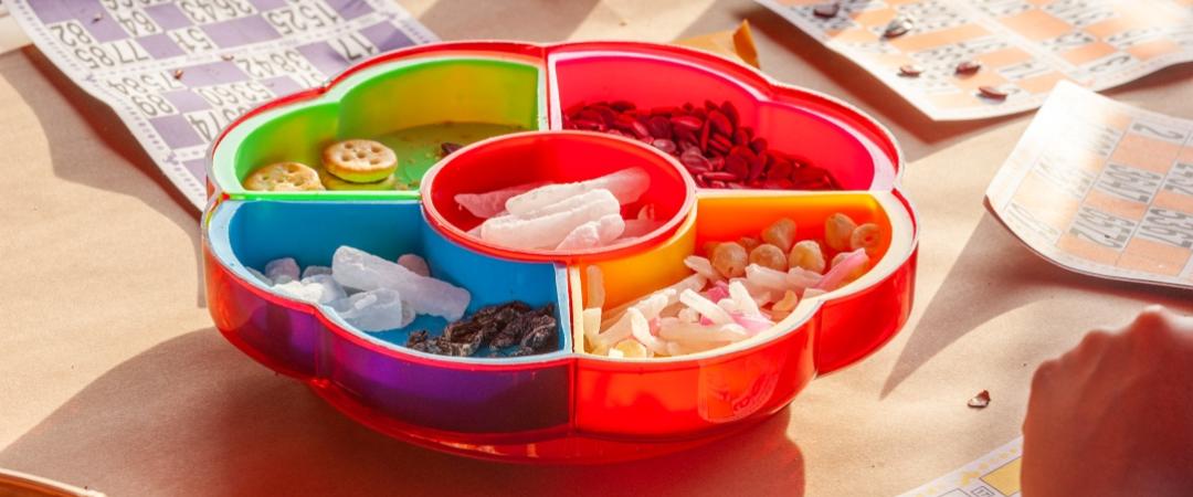 A colourful tray with assorted snacks is set on a table with bingo cards. Sunlight casts a warm glow, creating a playful, festive atmosphere.