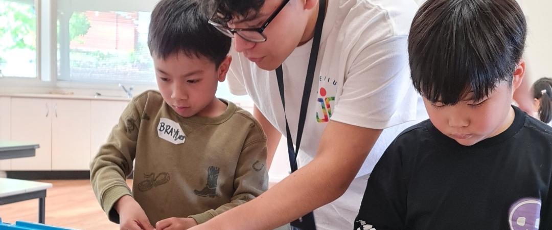 A man assists two boys with a robotics project, featuring colorful building blocks and gears. The indoor setting feels educational and collaborative.