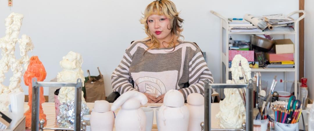 Artist seated at a cluttered desk with sculptures, surrounded by art supplies. The scene conveys creativity and focus in a studio setting.
