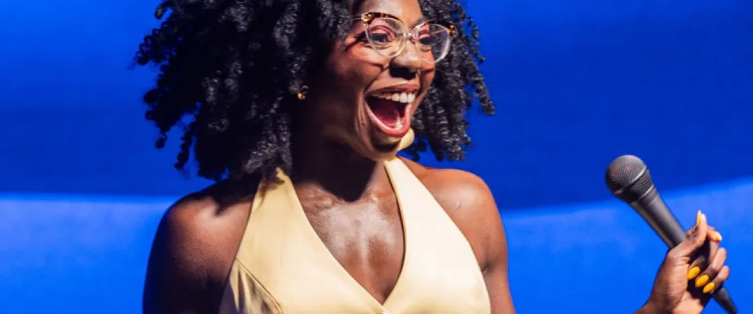 Smiling woman on stage holding a microphone, wearing glasses and a sleeveless beige top with buttons. She stands against a blurred blue background.