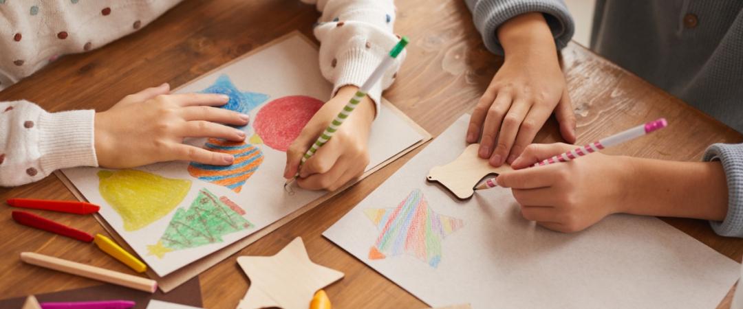 Children's hands are busy with colorful crayon drawings and star-shaped cutouts on a wooden table. The scene conveys creativity and playfulness.