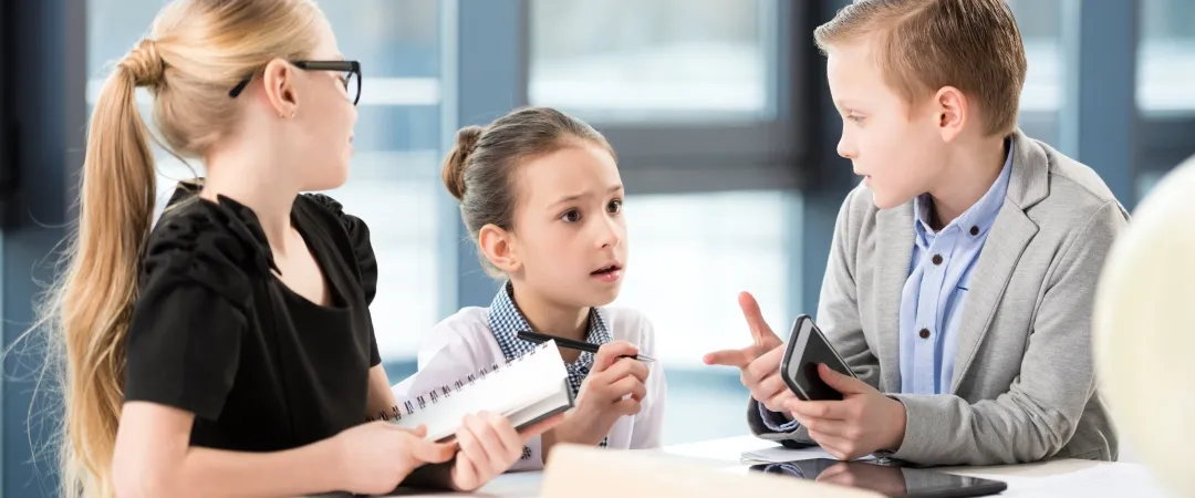 Two girls and a boy in officer clothes sitting at white desk with office supplies