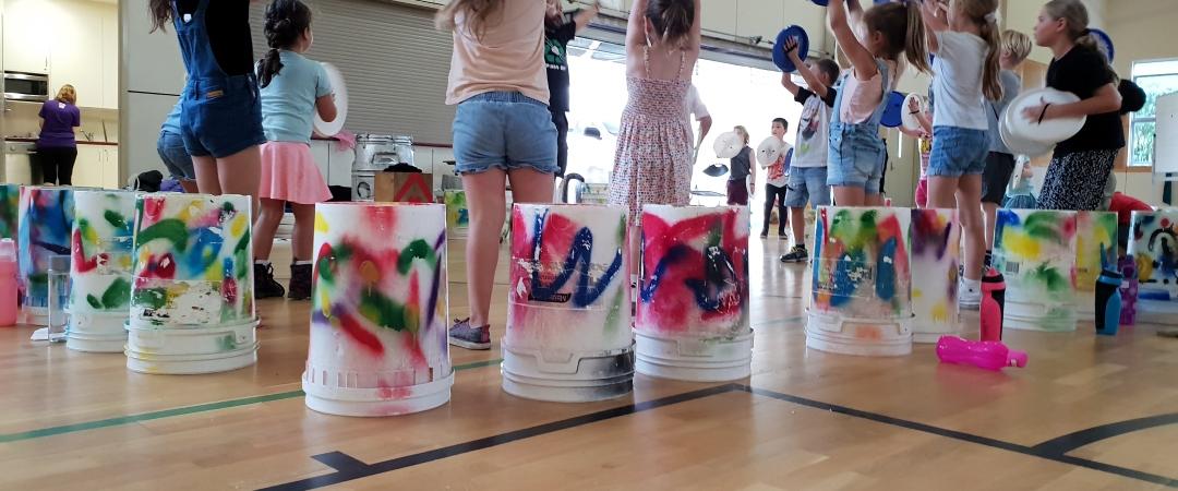 Children are inside a gym, each holding colorful buckets and plates, engaged in a lively drum circle. The mood is playful and energetic.