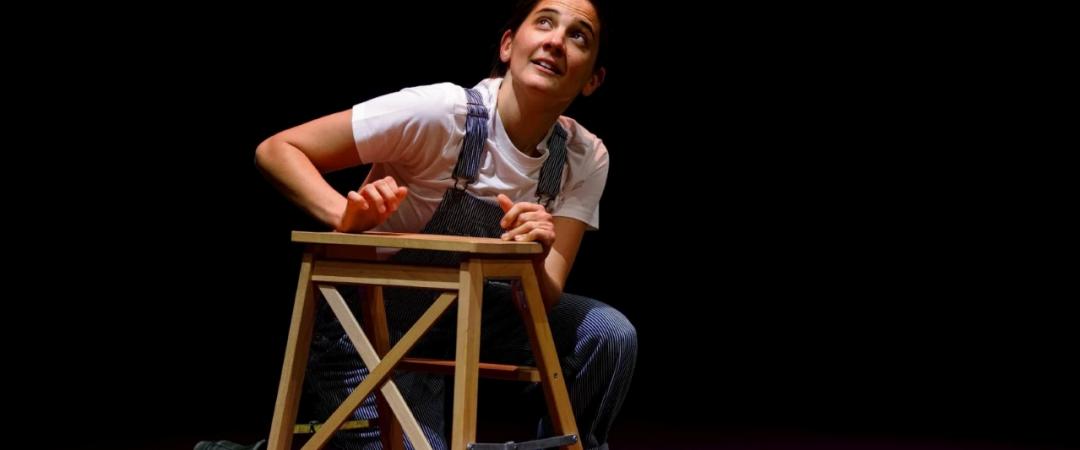 A person in overalls and a white shirt kneels beside a wooden stool on a colorful stage, looking upward with a thoughtful expression.