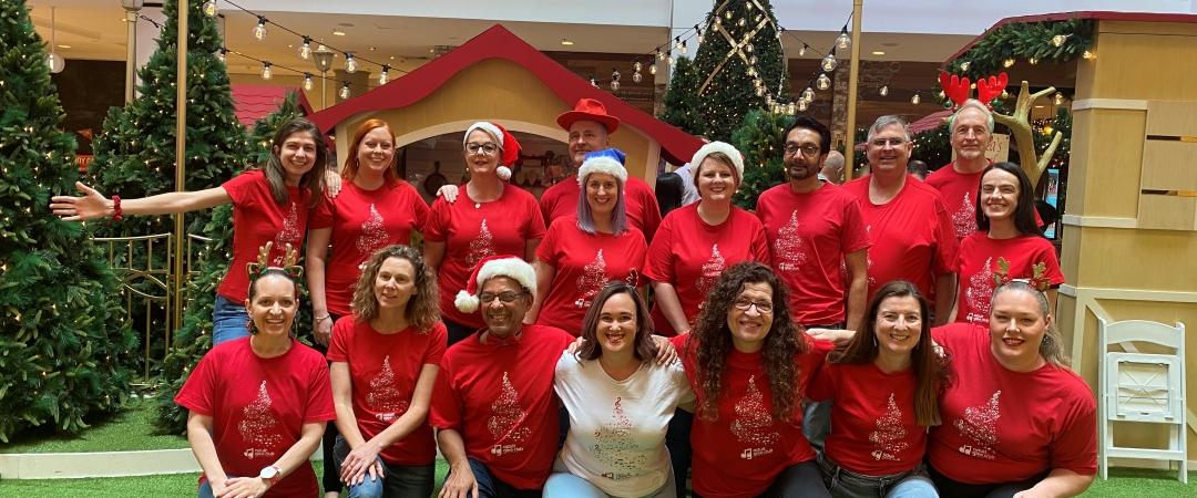 A group of smiling people, mostly in red shirts, pose in a festive setting with Christmas trees and decorations. Cheerful and joyful atmosphere.