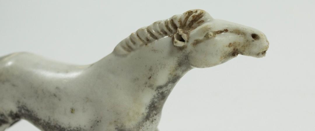 A small, antique ivory horse figurine with a weathered appearance. The textured mane and gentle curves evoke grace and history against a white backdrop.