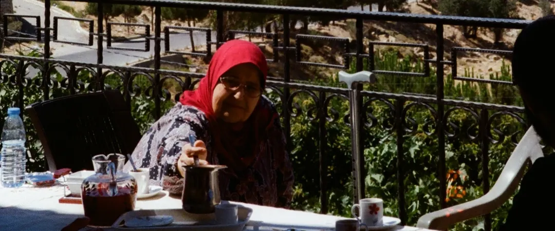 Woman in a red headscarf sits at a terrace table with tea, overlooking a rugged, sunlit valley; a companion sits nearby.
