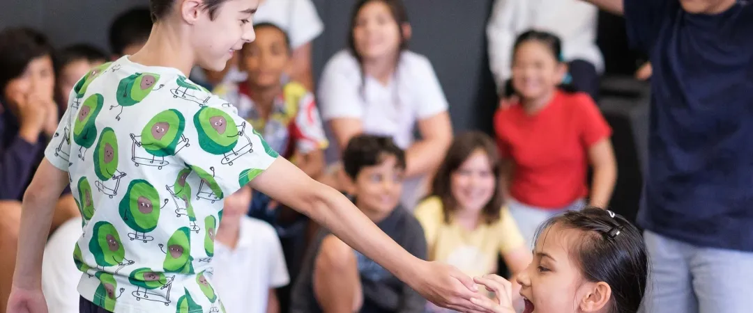 Young boy in an avocado-print shirt reaches toward a smiling girl as classmates watch and laugh during a lively group activity.