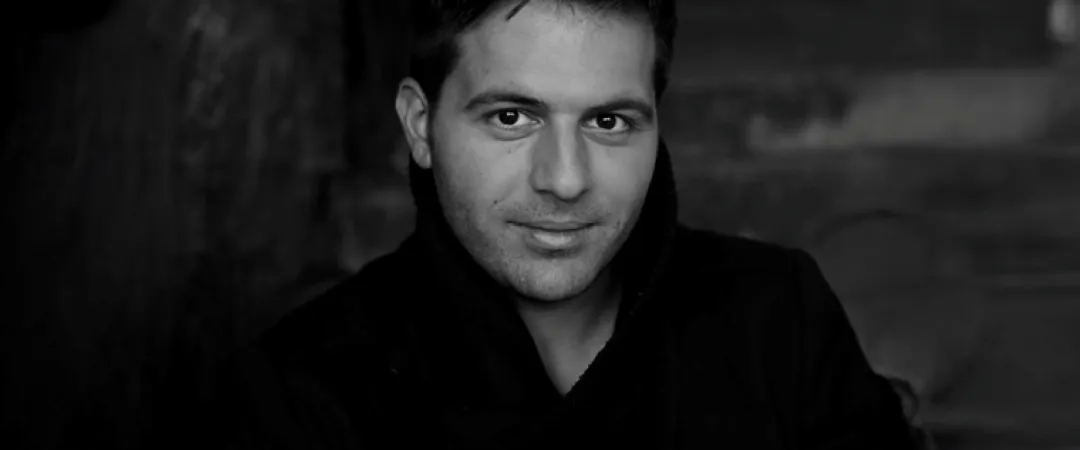 Black and white portrait of a man in a dark coat. He has short hair, a slight smile, and looks directly at the camera with a confident expression.