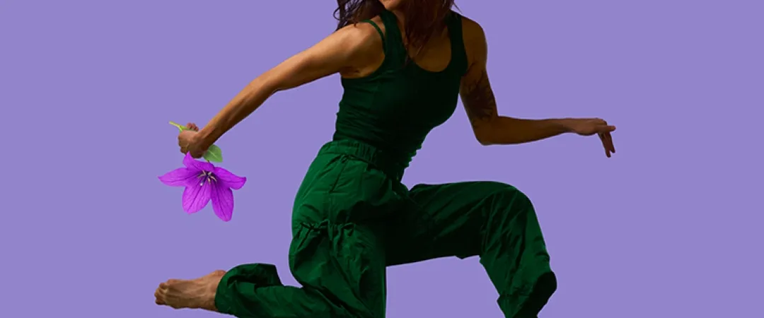 A woman in a green jumpsuit joyfully leaps with a purple flower in hand against a solid lavender background, conveying a sense of freedom and happiness.