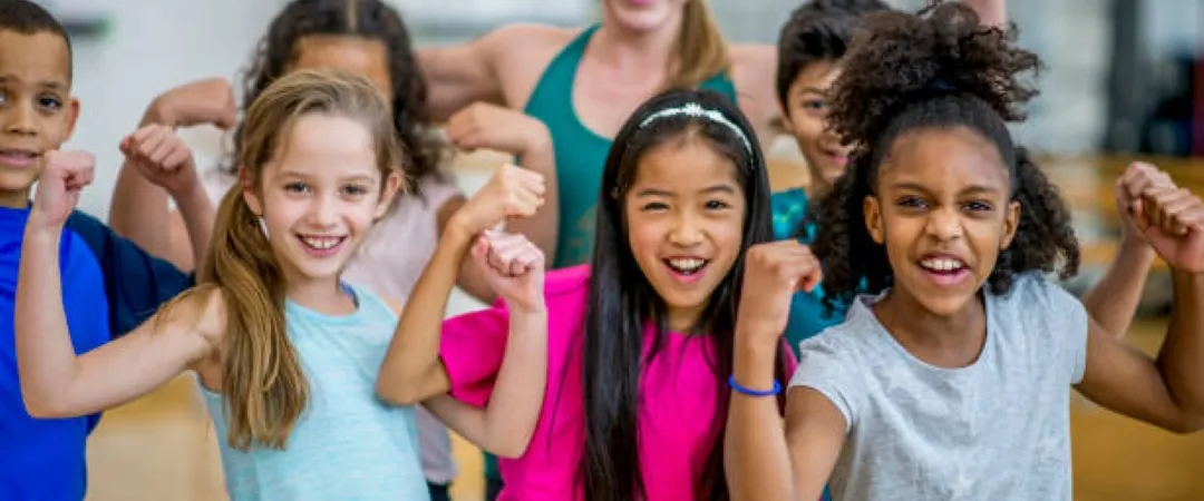 A joyful group of diverse children, along with a teacher, pose in a gym, flexing their arms and smiling enthusiastically, conveying energy and confidence.