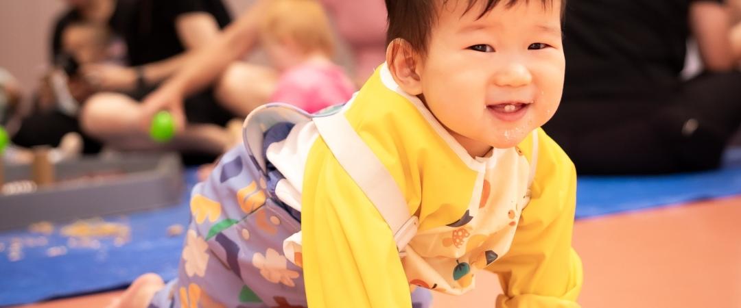 A smiling baby in a yellow outfit crawls on a play mat in a lively daycare setting. Other children and adults are blurred in the background, creating a warm, joyful atmosphere.