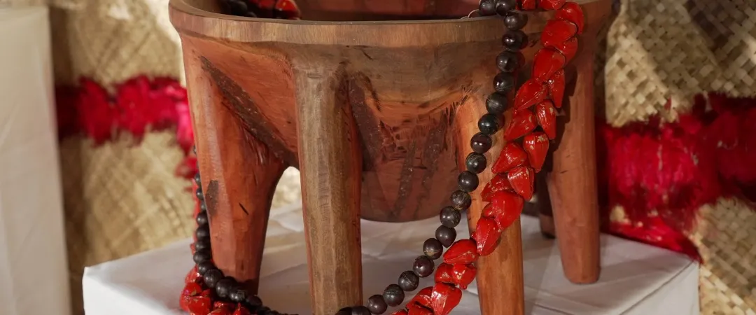 Carved wooden three-legged stool draped with red and black bead necklaces, displayed on a white-covered table against a warm woven backdrop.