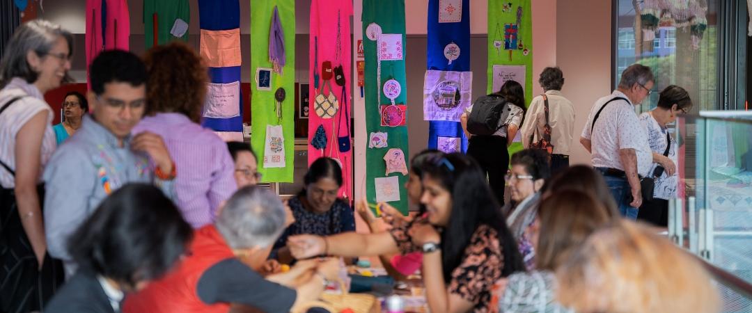 A vibrant community gathering, with people seated and interacting around tables. Colorful textile art pieces hang on the wall, creating an inviting, creative ambiance.