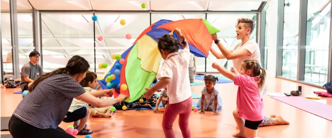 Children and adults play with a colourful parachute and scattered balls in a bright studio, creating a lively, joyful group activity.