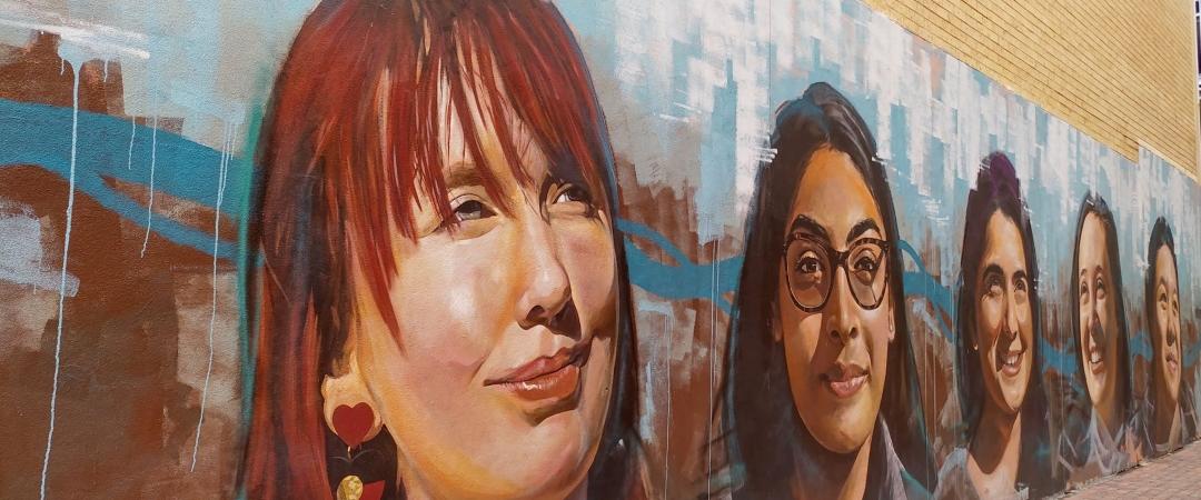 A vibrant mural on a brick wall depicts five smiling women of diverse backgrounds, set against abstract blue and brown brushstrokes. The atmosphere is uplifting.