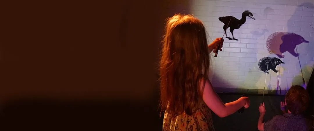 child standing holds emu shadow puppet, child sitting holding echidna puppet 