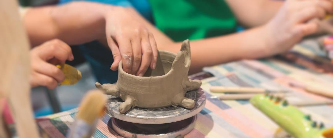 Children shaping clay into a bowl on a colorful tablecloth, surrounded by brushes and tools. The scene conveys creativity and focus in a pottery class.