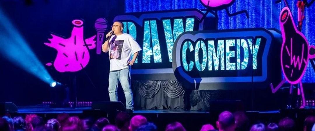 A comedian performs on stage under bright blue and purple lighting, with a large ""Raw Comedy" sign. The audience is visible, and laughter fills the lively atmosphere.