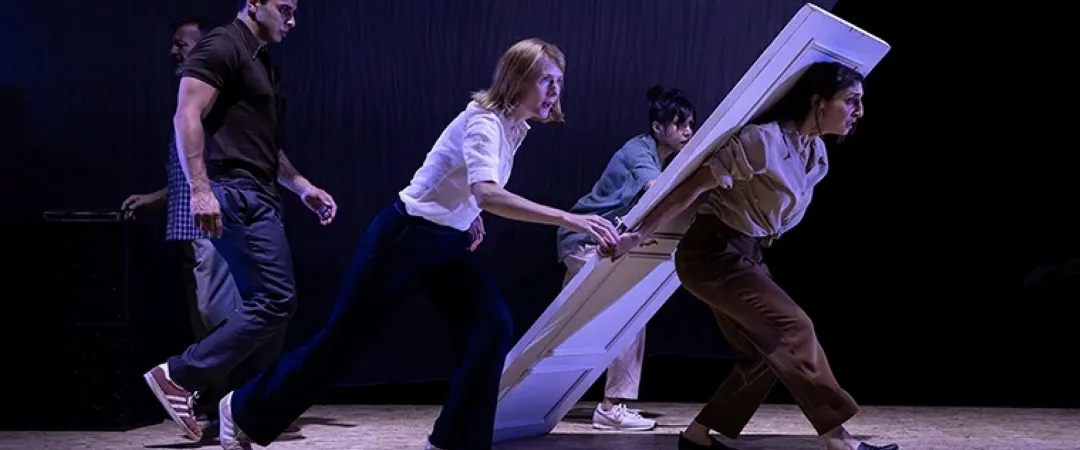 On a dimly lit theatre stage, five performers push and pull a tilted white frame, creating a tense, energetic contemporary dance scene.