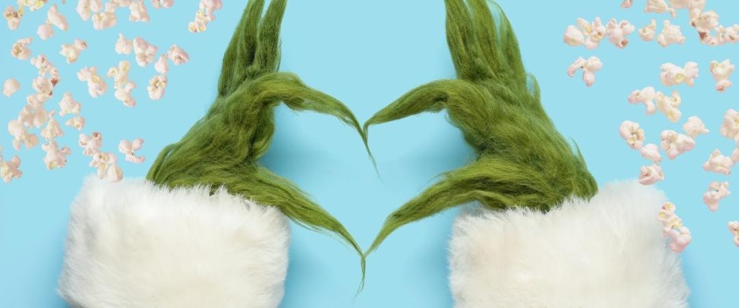 Two furry green hands with long fingers forming a heart shape against a blue background, surrounded by scattered popcorn. The scene feels playful and whimsical.