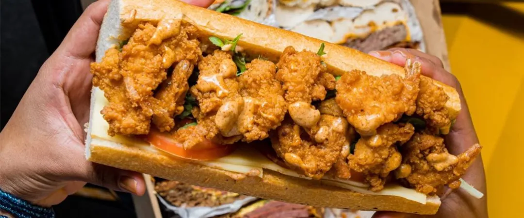 Hands hold an overstuffed fried chicken sandwich with crispy pieces, sauce, lettuce, and tomato; assorted sandwiches fill the background.