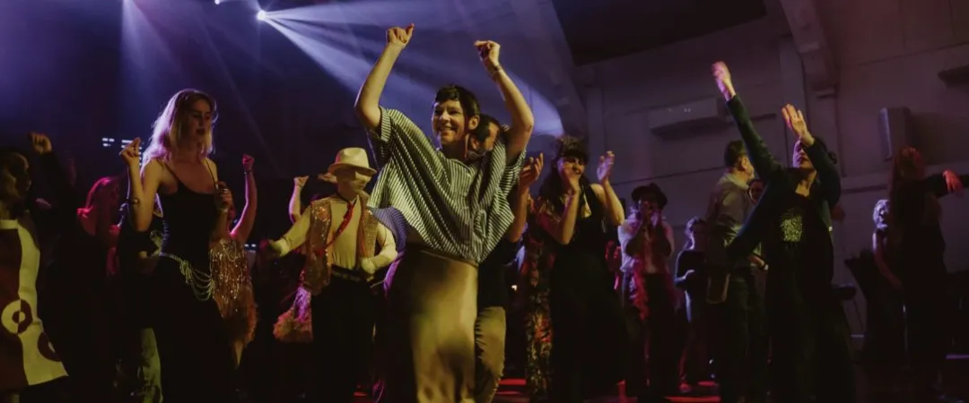 A lively crowd dances at a concert under purple stage lights, led by a smiling person with both arms raised; the scene feels joyful and energetic.