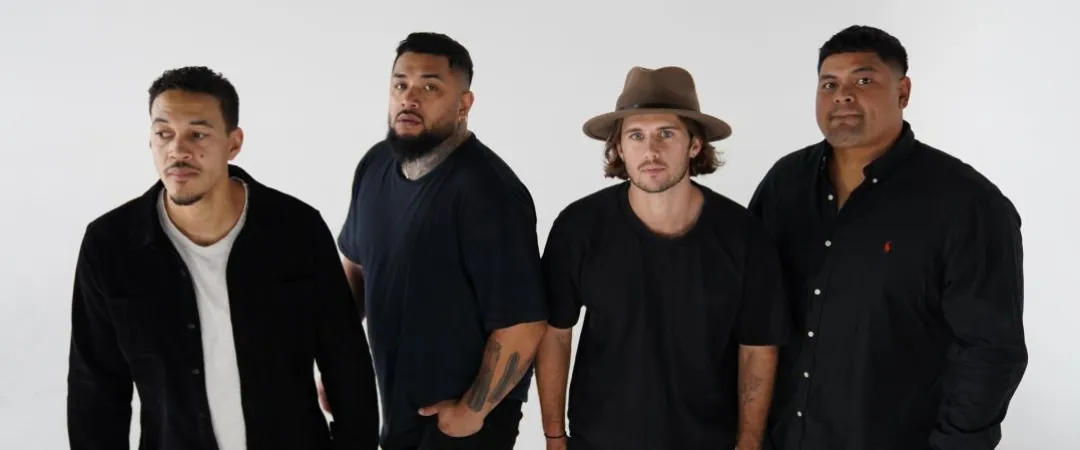 Four men stand against a white background, looking serious. They wear casual black outfits; one wears a hat. The atmosphere is confident and focused.