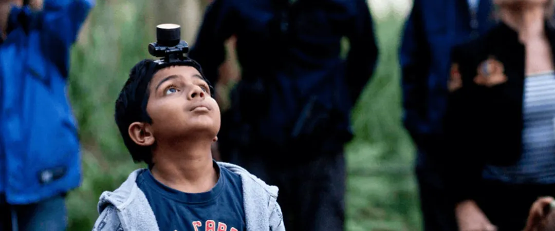 Young boy with a small camera mounted on his head looks upward in a forest, surrounded by blurred adults; curious, playful mood.