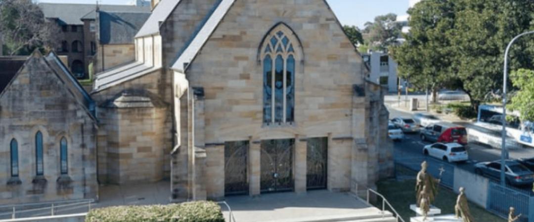 A historic stone church with arched windows, tall steeple, and steps leading to the entrance. Statues stand on the right, with trees and cars nearby.