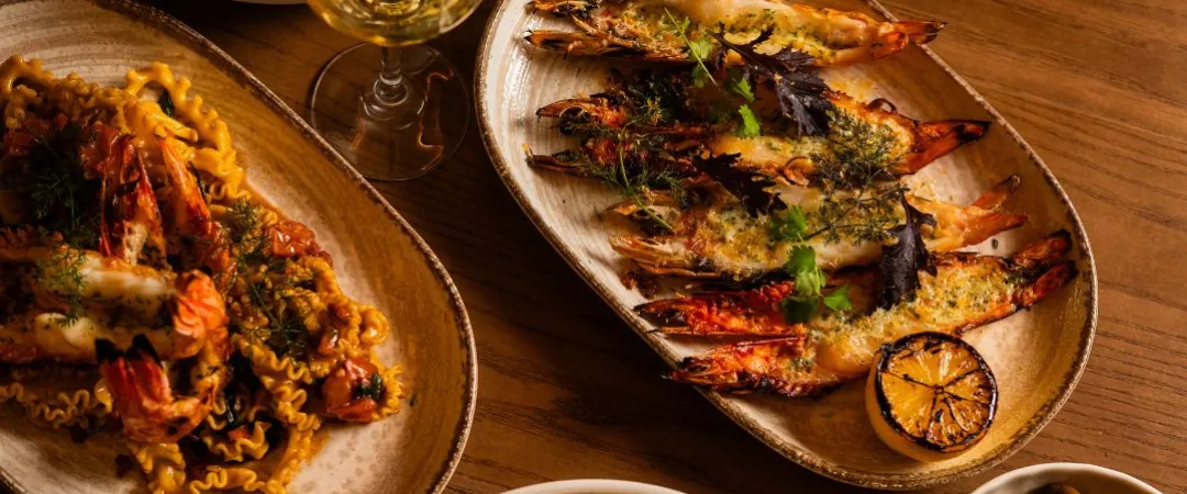 A warm-toned table setting with grilled shrimp, pasta, and edamame bowls. A glass of white wine adds elegance, while herbs enhance a gourmet feel.