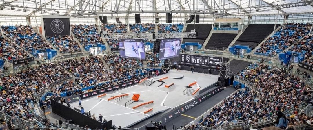 A packed arena with a transparent roof hosts a skateboard competition. Enthusiastic fans fill the seats. Two large screens display live game action.