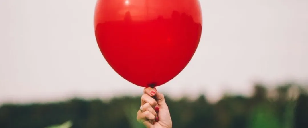 Close-up of a hand holding a bright red balloon outdoors against a softly blurred background of greenery and sky.