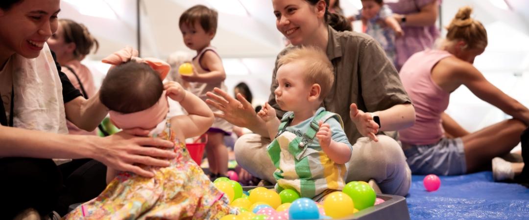 Babies play in a colorful ball pit on a blue mat, surrounded by smiling adults. The scene is lively and joyful, with a focus on family interaction.