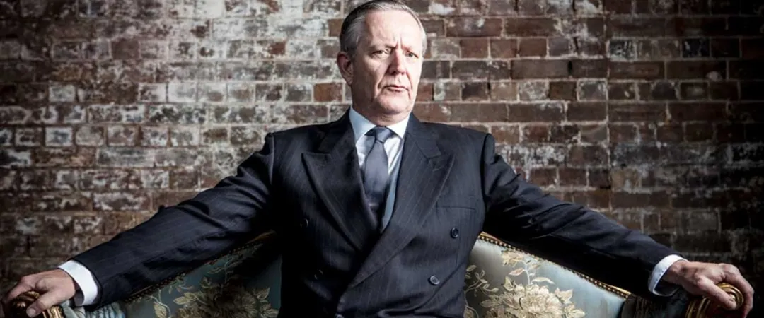 A man in a suit is seated on a couch, looking relaxed and engaged in thought.