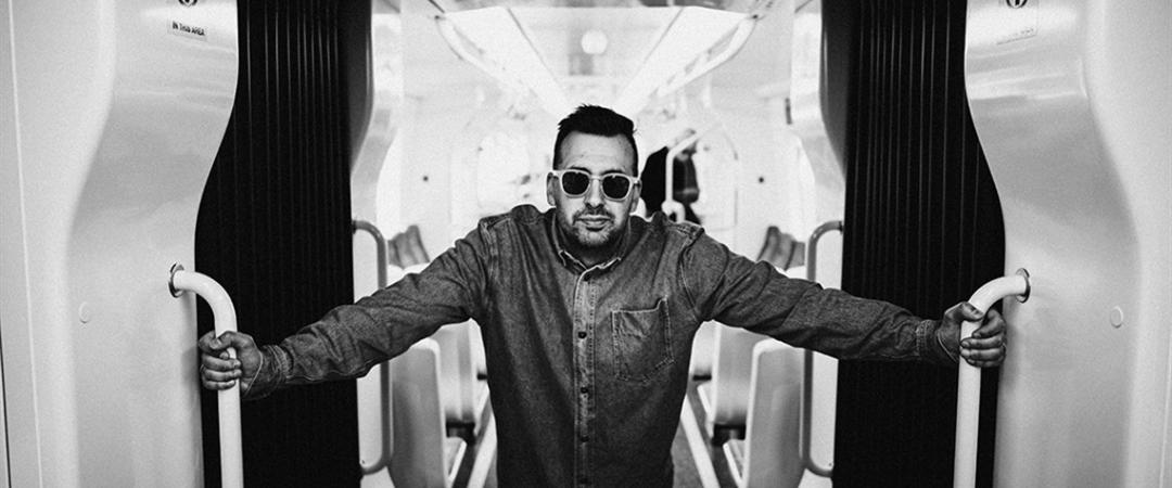 Black and white image of a person wearing sunglasses and a jacket, leaning casually with arms outstretched on a narrow walkway, conveying confidence.