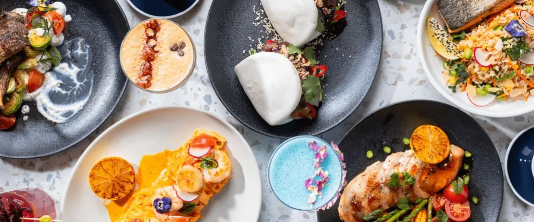 Grey table with plates of food like bao buns, salmon & rice, pasta, chicken and corn ribs. 