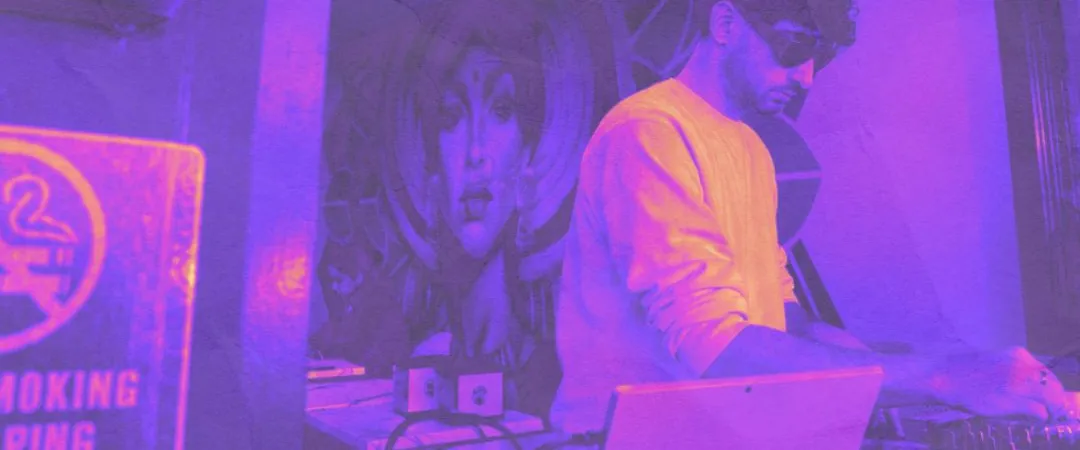 A focused DJ in an orange shirt works a turntable beside an open laptop, bathed in purple-pink light, with a large portrait mural behind him.