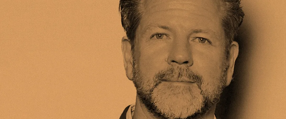 Sepia-toned head-and-shoulders portrait of a bearded man in a dark jacket, gazing calmly at the camera against a plain warm background.