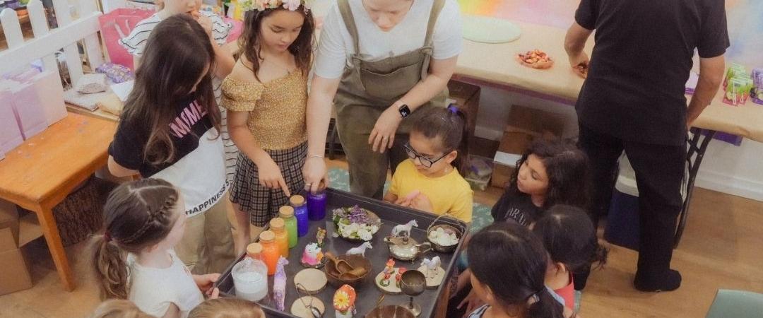 A group of children and two adults gather around a table with art supplies in a classroom. A colourful rainbow decorates the wall, creating a lively atmosphere.