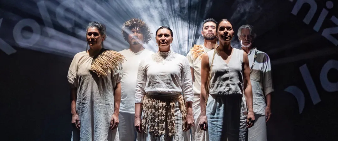 A group of six performers stands confidently on a dark stage, dressed in white, rustic outfits. Dramatic lighting casts shadows and highlights their expressions. Text projections on the backdrop create a dynamic atmosphere.