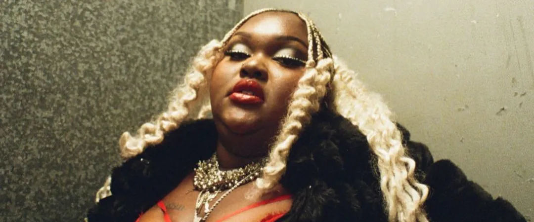 A woman with blonde braids and bold makeup, wearing silver jewellery and a black fur coat over a red top. She poses confidently, conveying power and elegance.