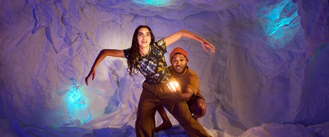 A woman poses dynamically in a tattered cave-like setting with blue and purple lighting, while a man behind her shines a light, adding a sense of mystery.
