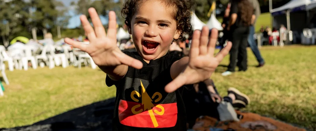 A child with curly hair smiles widely, playfully displaying open palms in a park. Green grass, trees, and people milling around under white tents provide a vibrant, joyful outdoor setting.