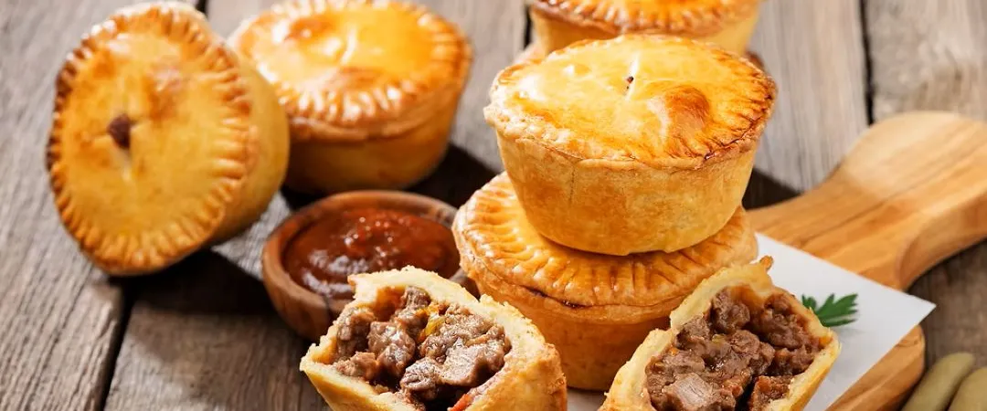 Golden meat pies sit on a wooden board, some cut open to reveal savory beef filling, with dipping sauce nearby; warm, rustic, appetizing.