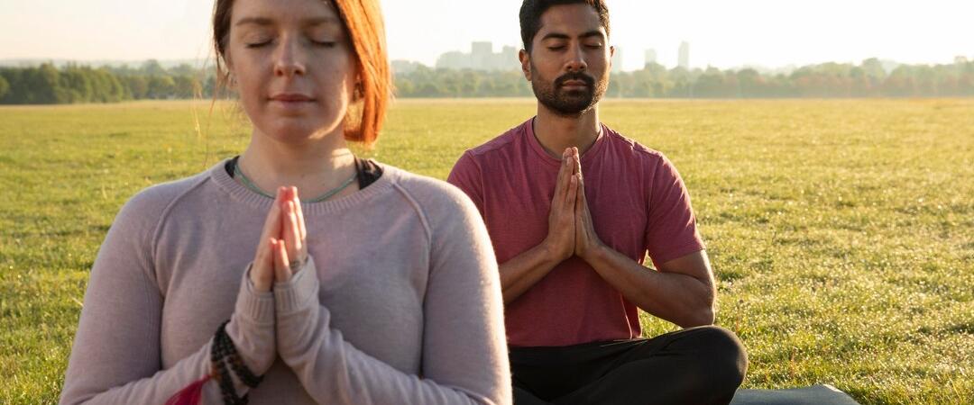 A man and a woman practice yoga outdoors on a sunny day, sitting cross-legged with hands in prayer position, conveying calm and focus in a grassy field.