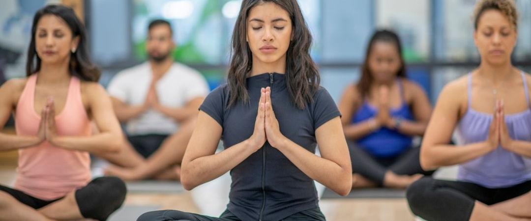 A diverse group of people in a yoga class sit cross-legged on mats, eyes closed, hands in prayer position. The setting is calm and focused.