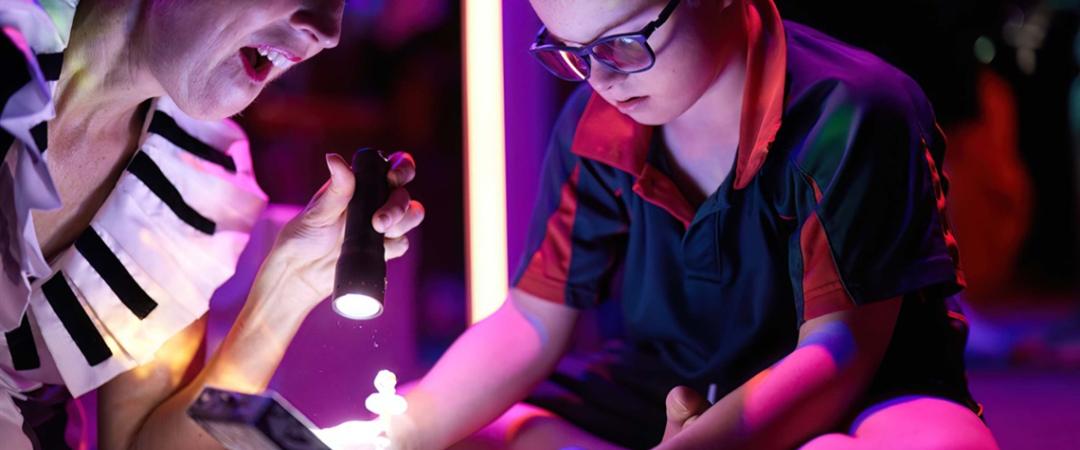 A person in a theatrical costume, excitedly holding a flashlight, illuminates an open box that a young boy with glasses is curiously examining. The setting is vibrant, magical.